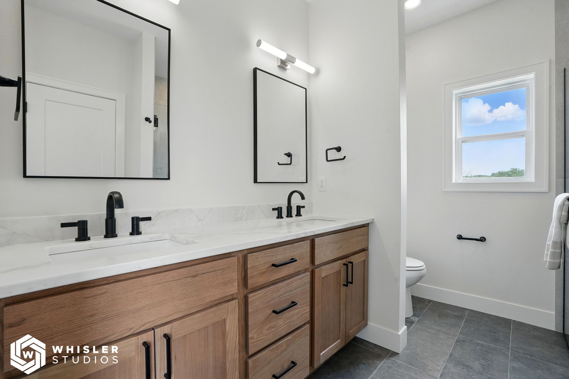 A bathroom with two sinks, two mirrors, a toilet and a window.