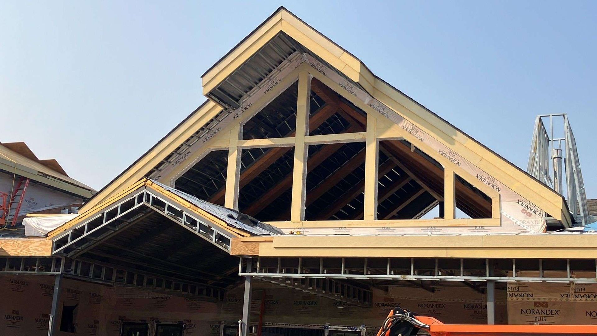 Construction site with wooden framing for a roof and window, metal beams, and an orange excavator.
