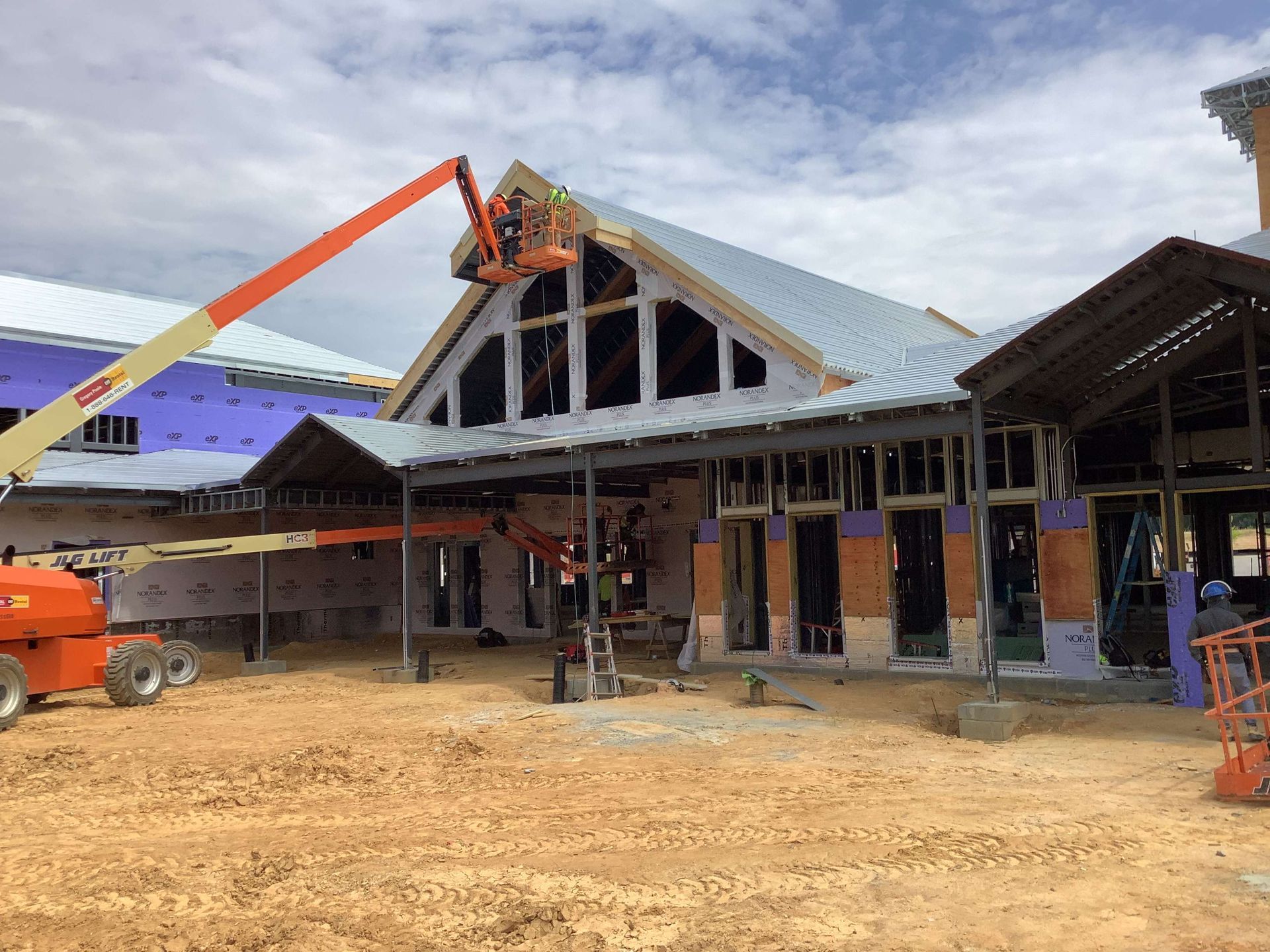 Construction site: metal roof installed on a building with large windows. An orange lift is in use.