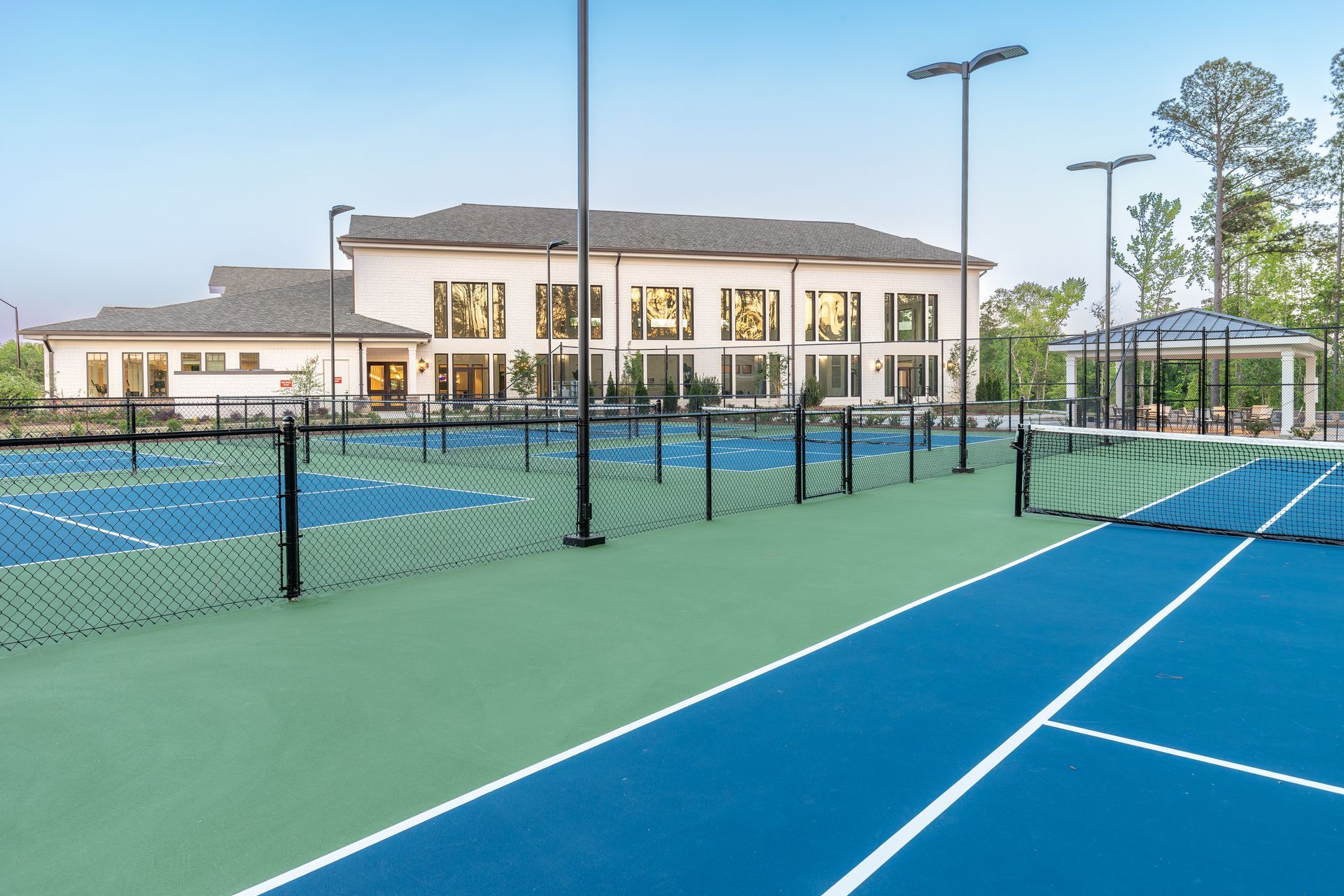 Tennis courts with a building and gazebo in the background. Blue and green courts, black nets, and lampposts.