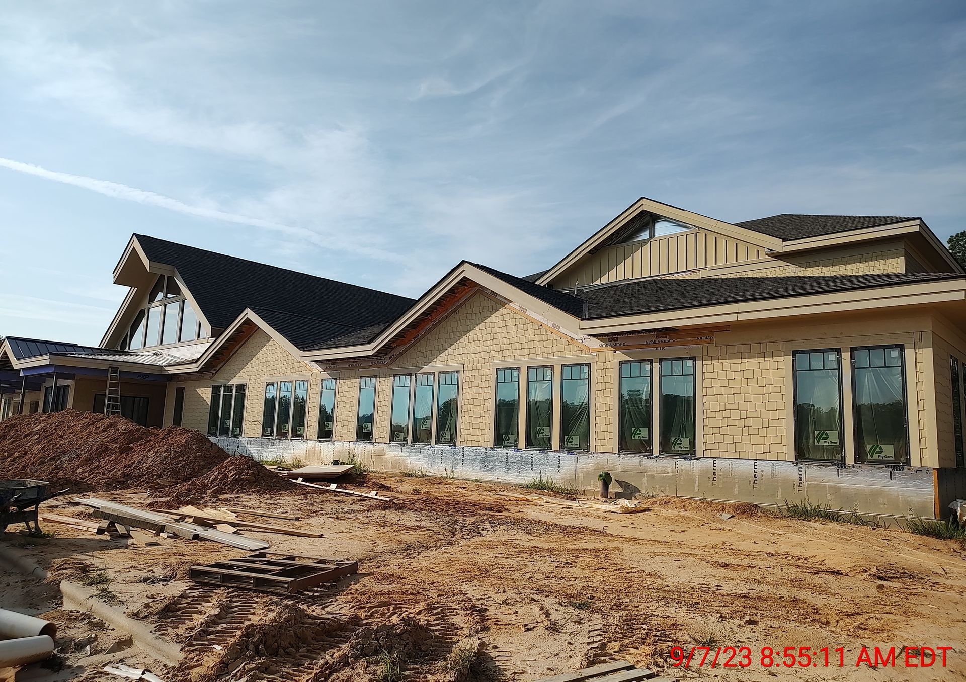 Construction site: unfinished building with multiple windows, brown siding, and dirt ground under a blue sky.