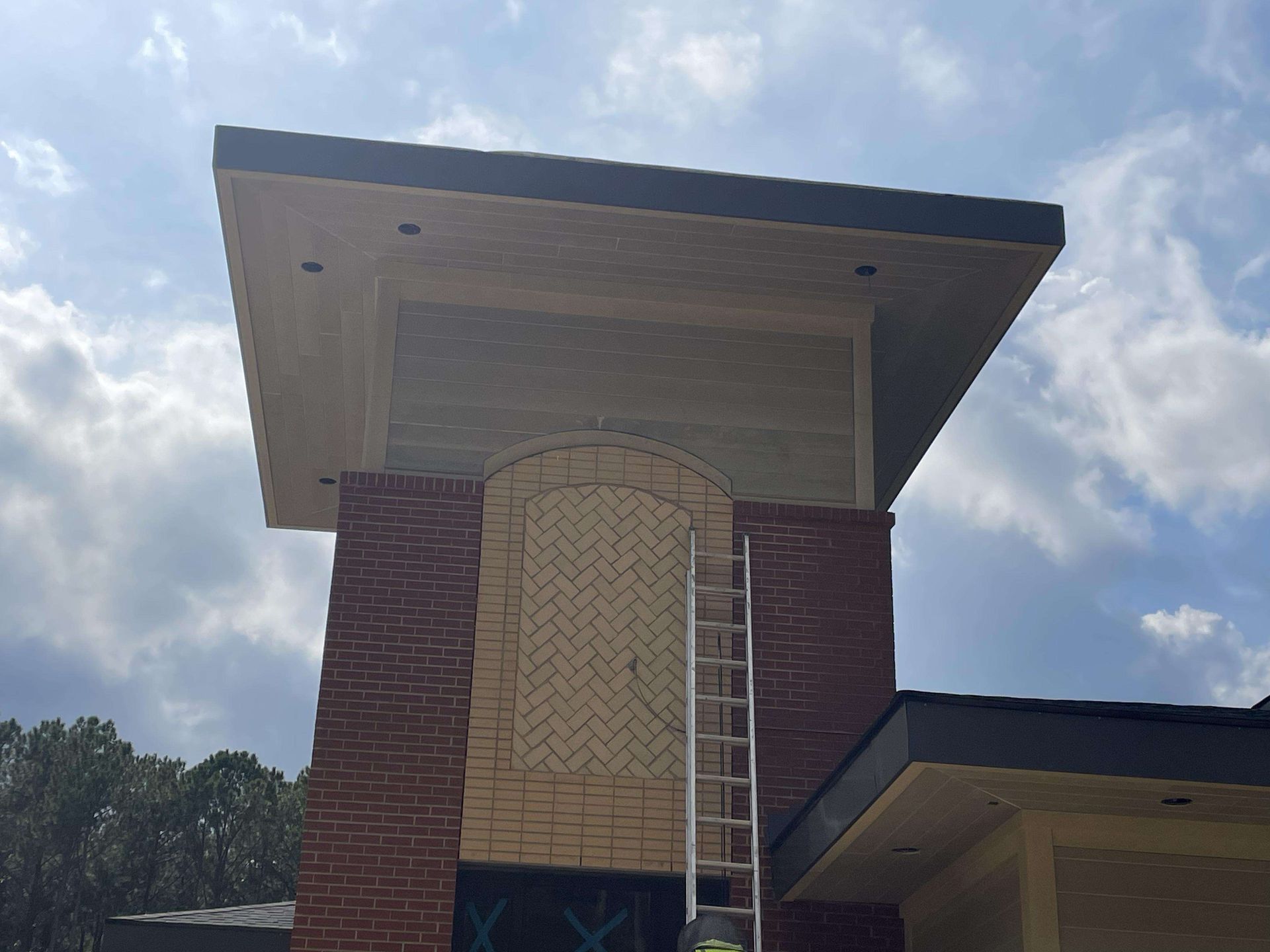 Brick building with a decorative arched panel, a ladder, and a dark roof against a cloudy sky.