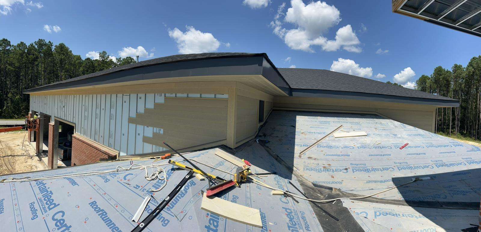 House under construction, partially covered with shingles and siding, surrounded by trees, blue sky with clouds.