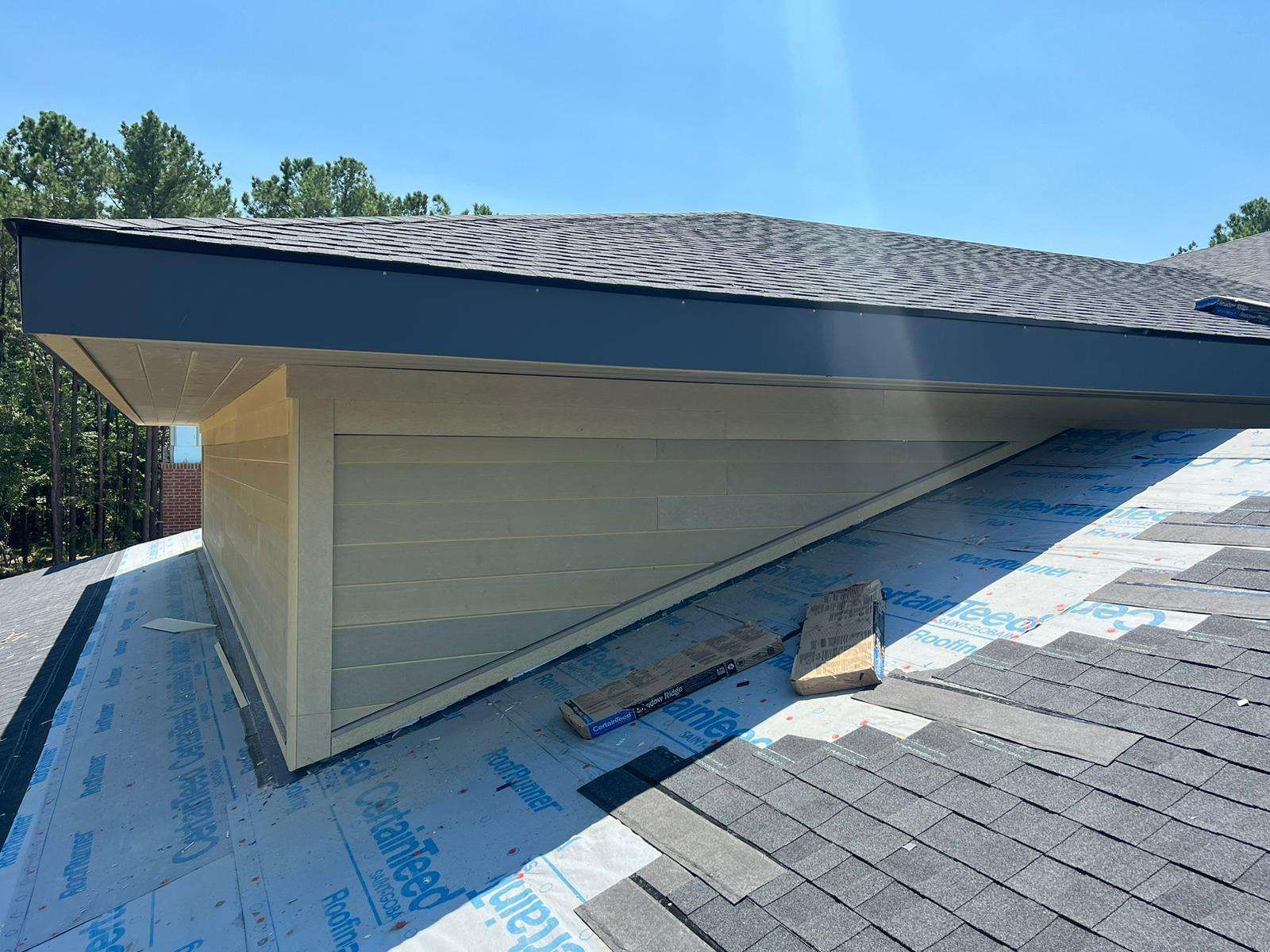 Close-up view of a roof with shingles, light siding and dark fascia under a clear blue sky.