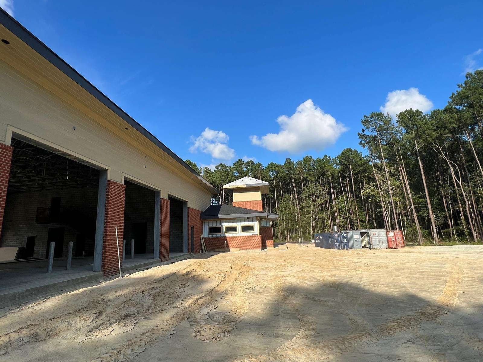 Construction site: a brick building with open garage doors, a watch tower, and trees under a blue sky.