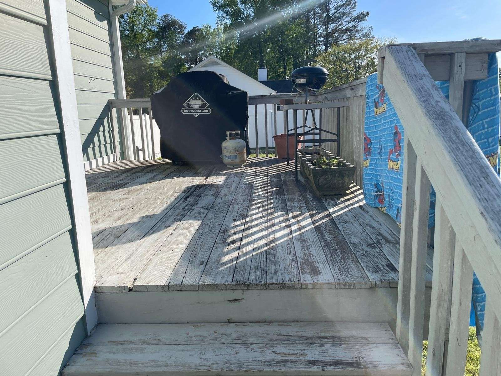 Wooden deck with weathered boards and railing; grill under a cover, a gas tank, and foliage are visible.
