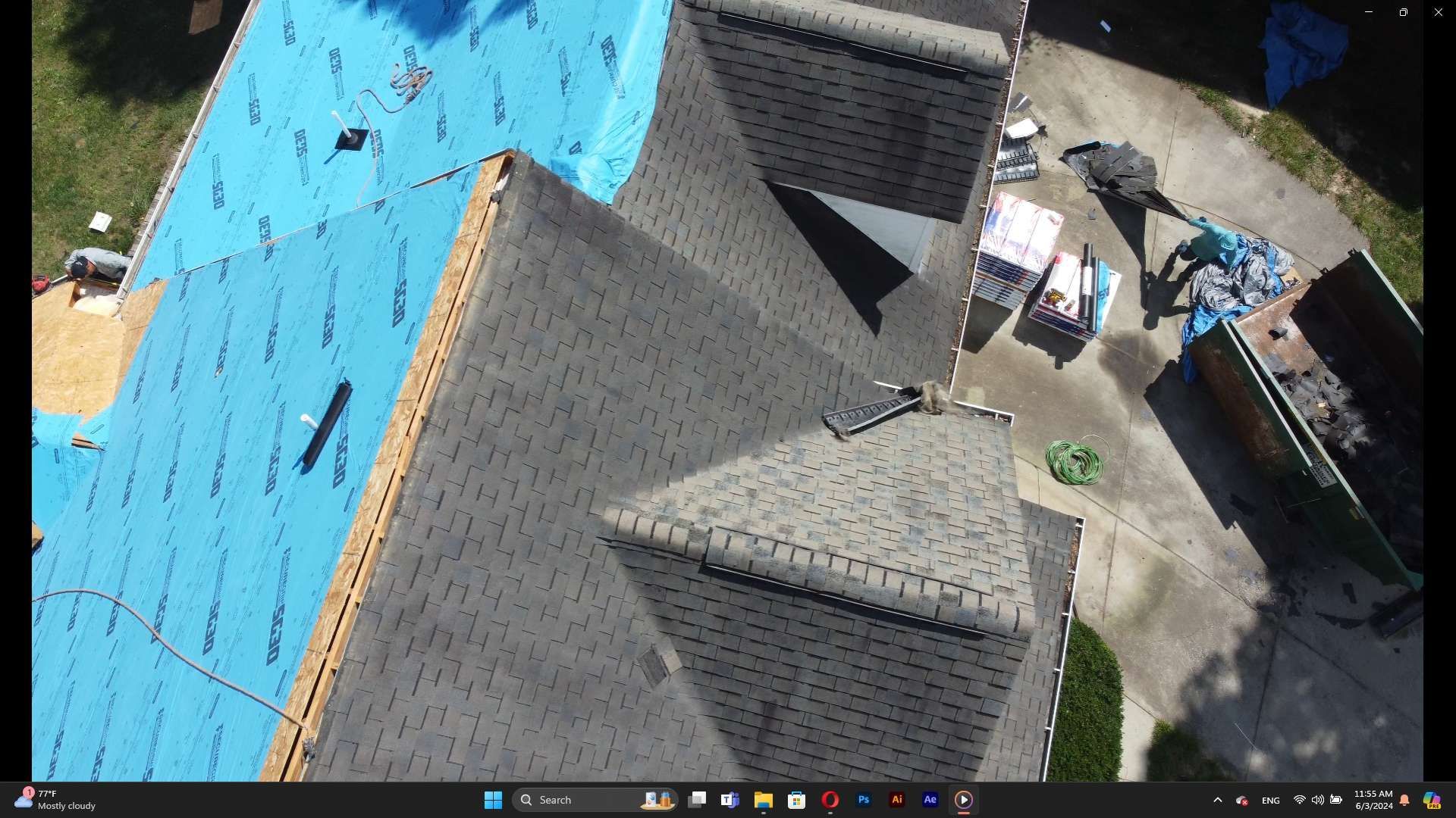 Aerial view of a house with a roof under construction. Blue underlayment, gray shingles, debris, and a dumpster are visible.