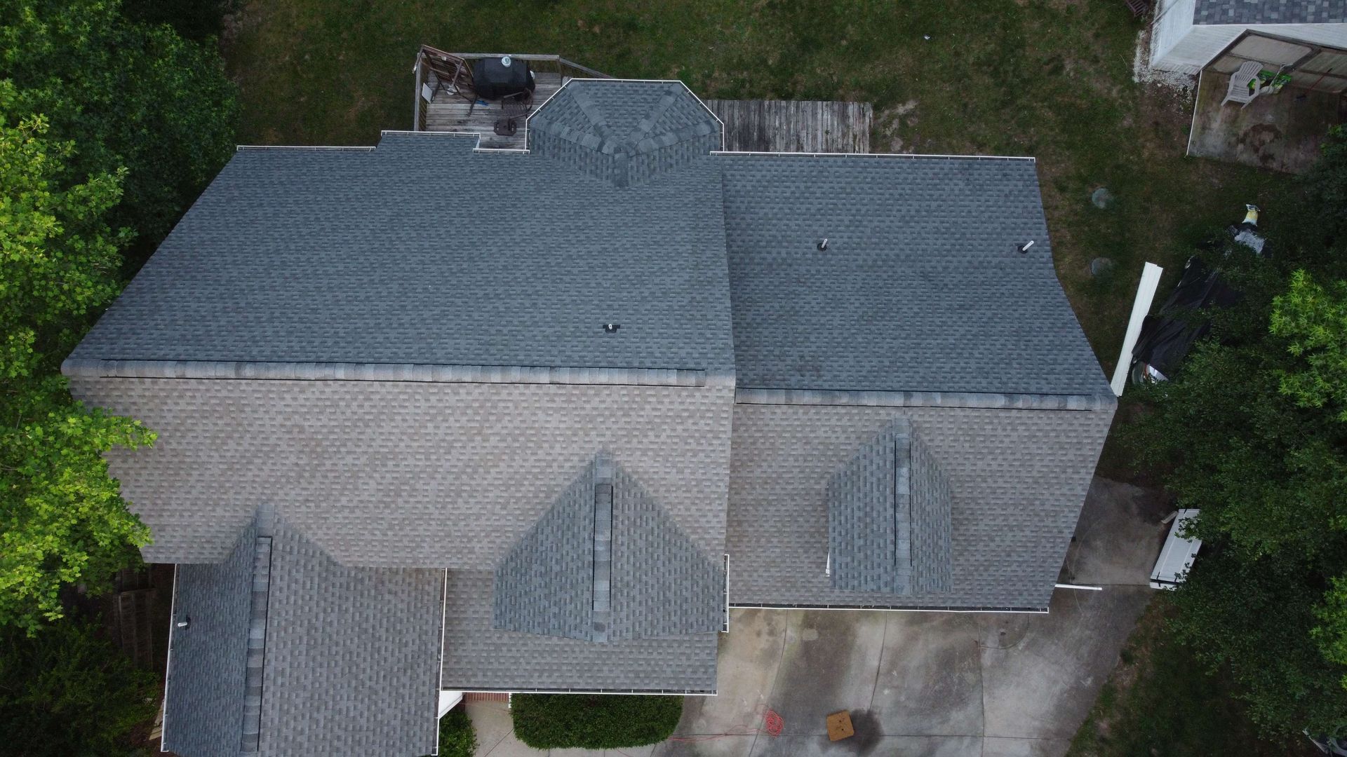 Overhead view of a gray-roofed house with a complex roofline, surrounded by trees and a concrete driveway.