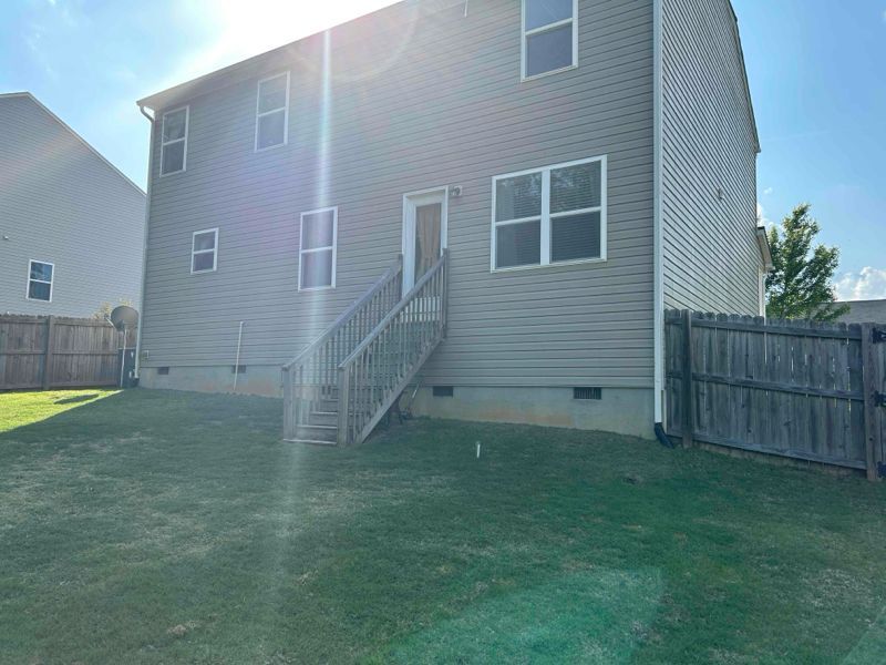 Back of a two-story house with gray siding, wooden stairs to a back door, and a green lawn, sunny day.