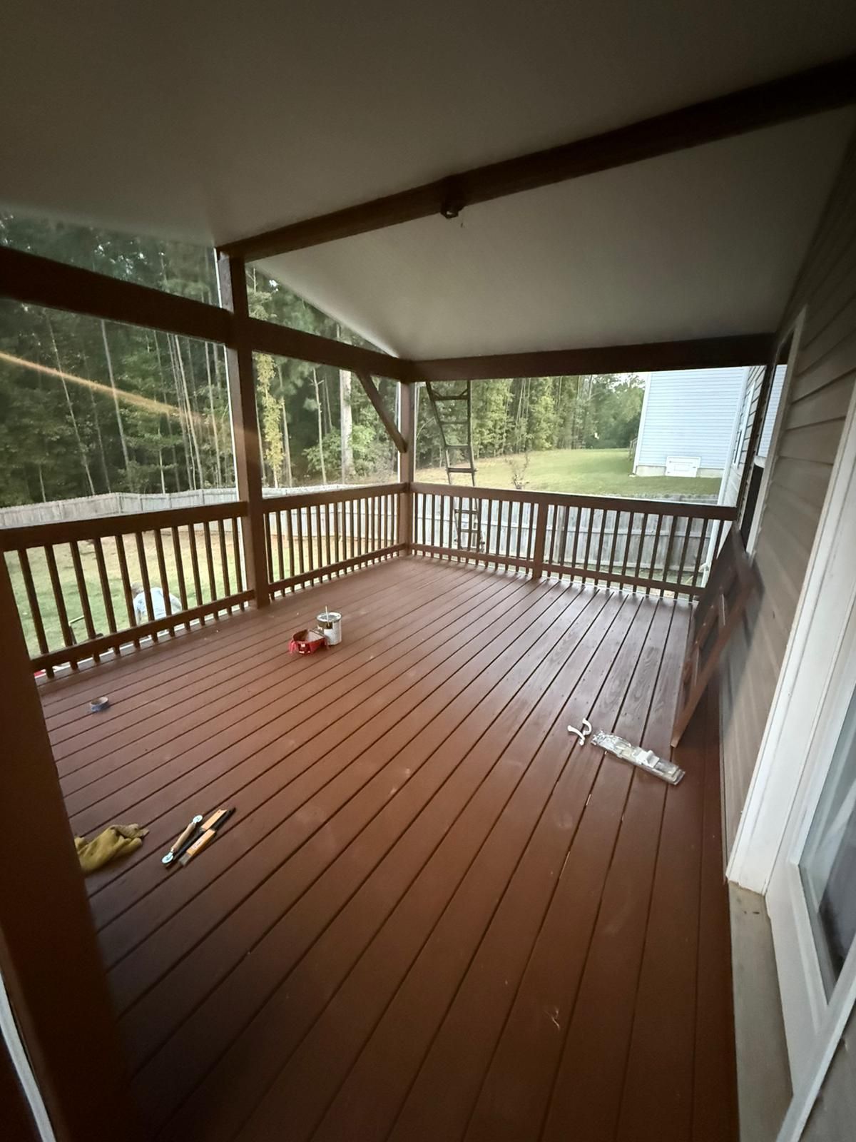 Covered wooden deck with railing, overlooking a wooded area. Brown stain on deck and structure.