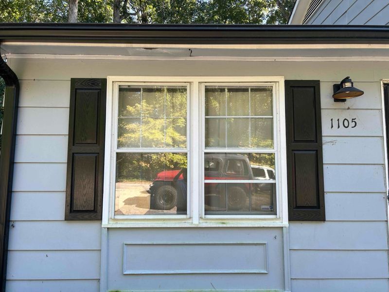 Light blue house with white-framed double window, black shutters, and address 1105. A red vehicle is reflected in the glass.