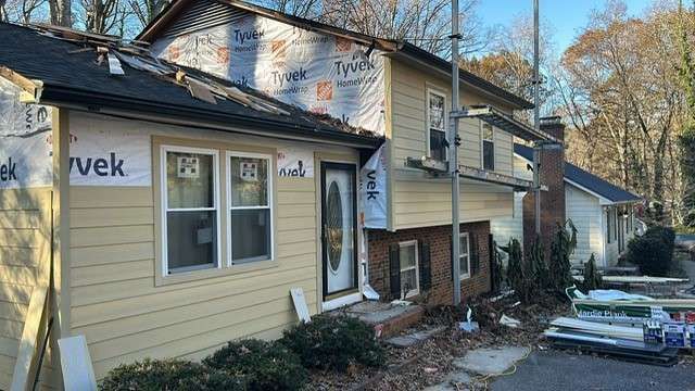 House under construction, covered in Tyvek. Scaffolding and materials nearby. Yellow siding, black door, trees in the background.
