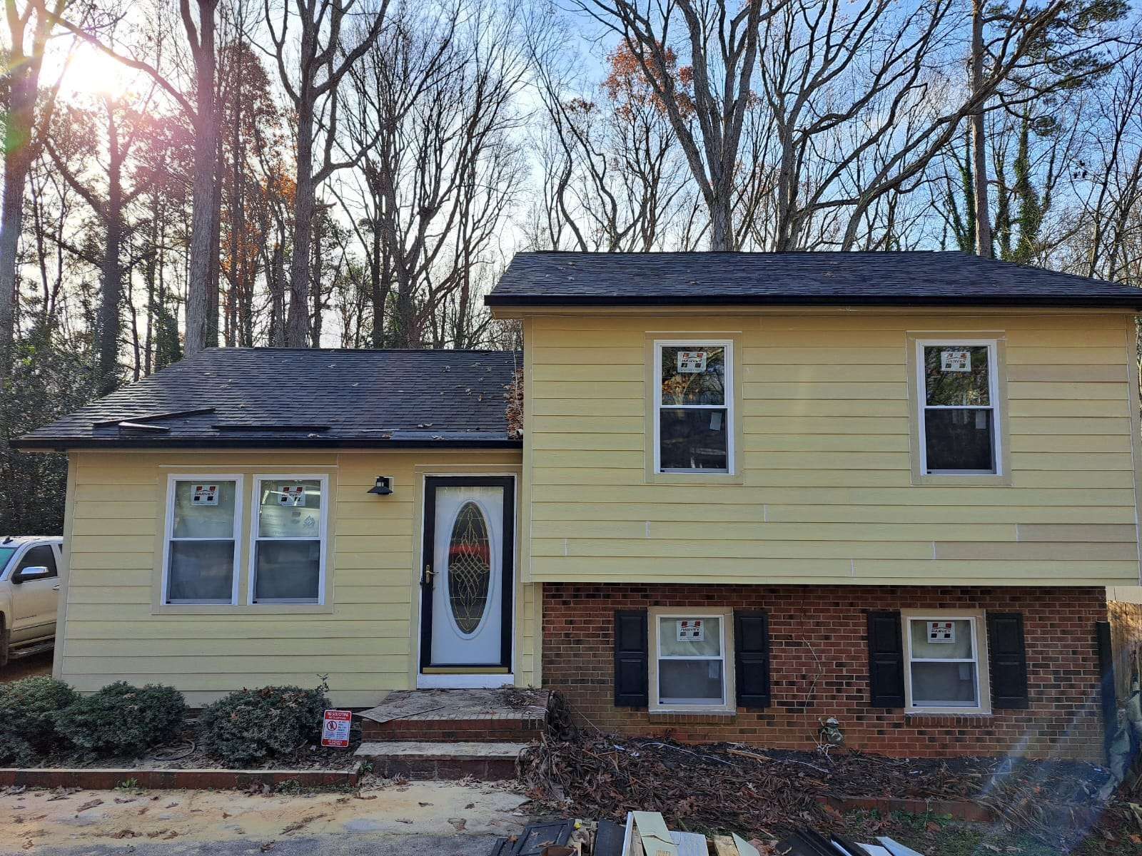 Yellow house with brick facade, windows, and damaged roof, set against trees.