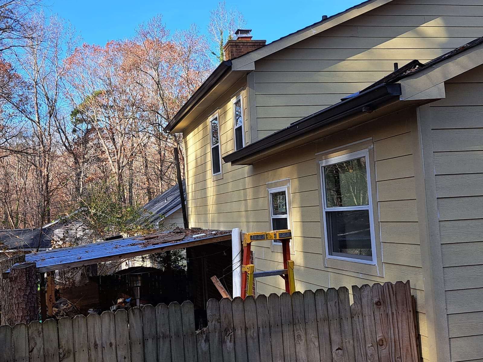 Yellow house exterior with brown roof, fence, and leafless trees in background.