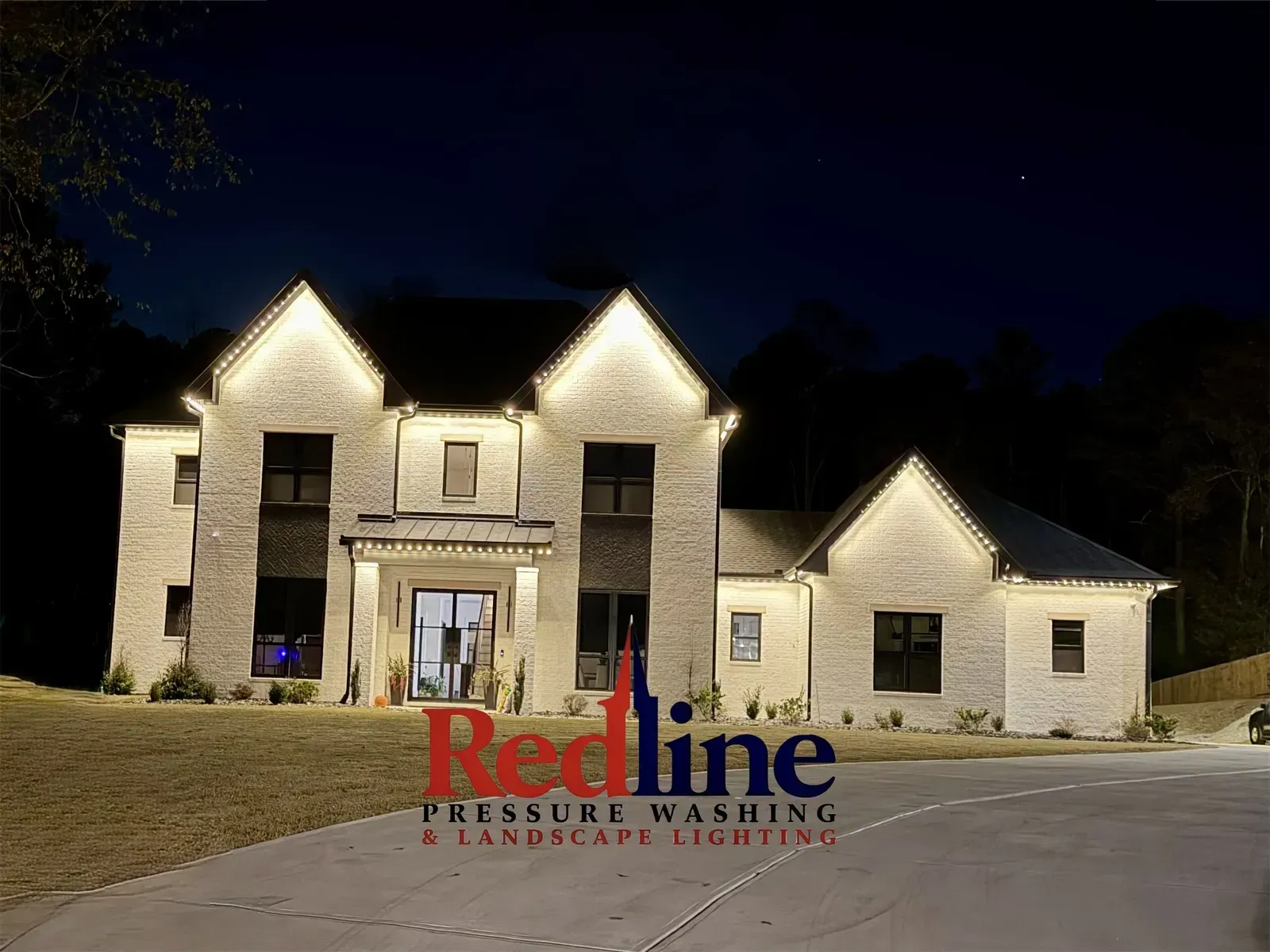 Illuminated stone house at night with a Redline Pressure Washing logo over the driveway