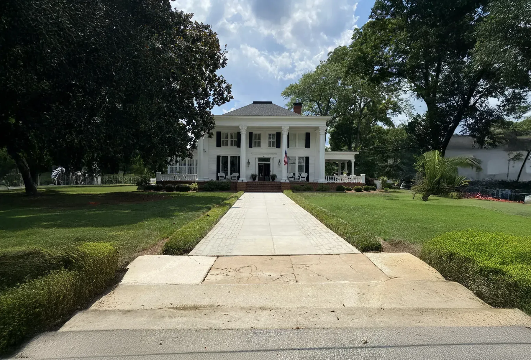 White house with large front porch and long concrete walkway, surrounded by green lawn and trees.