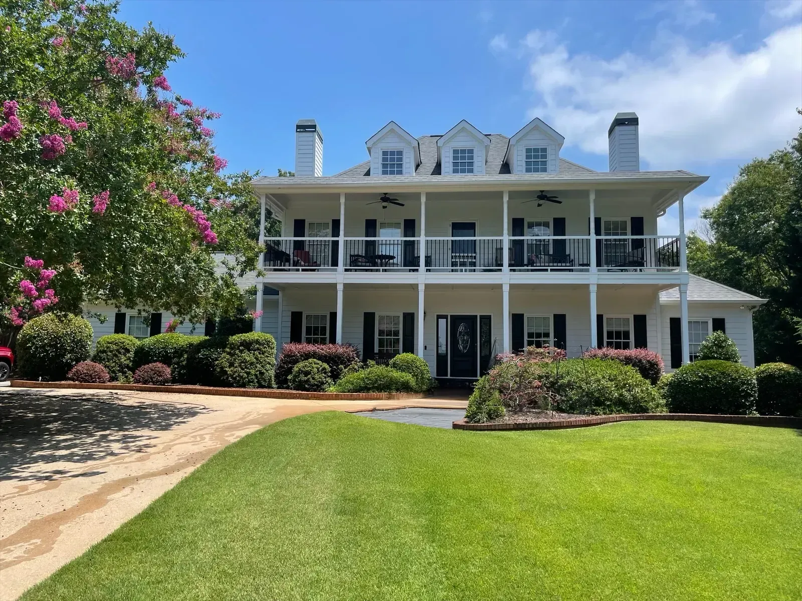 Large white two-story house with wraparound porch, manicured lawn, and blooming pink tree under a blue sky