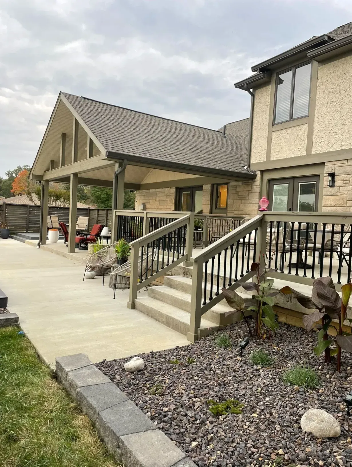 A stone home with a covered patio, wooden deck, stairs, and a decorative rock garden under a cloudy sky.