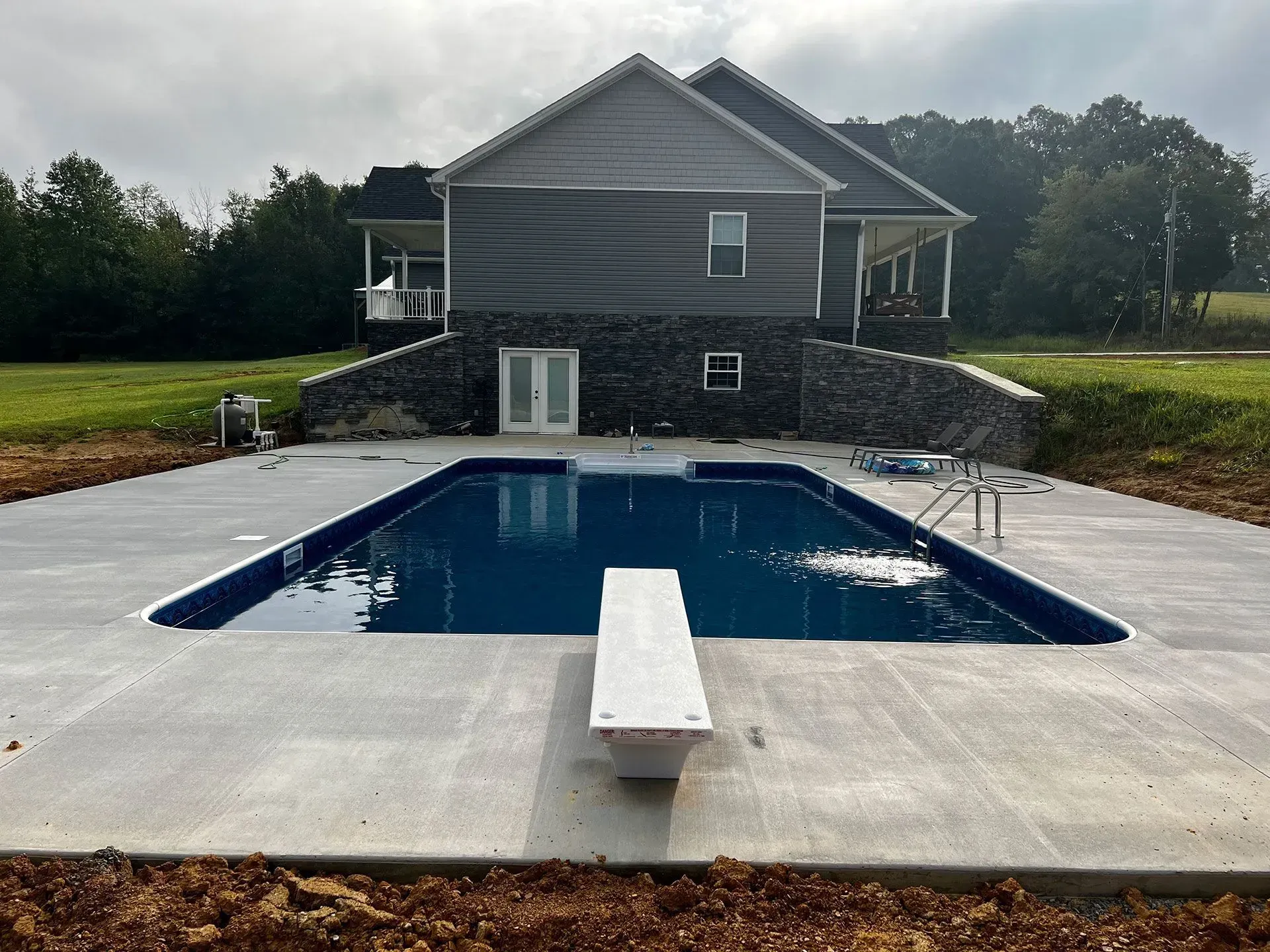 Rectangular swimming pool in front of a gray house. White diving board, concrete deck.