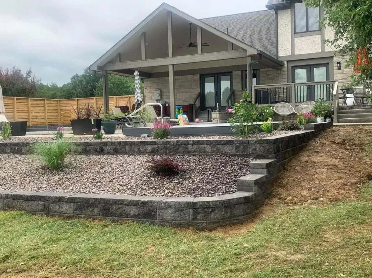 Backyard with tiered retaining walls, patio, and house under a cloudy sky.