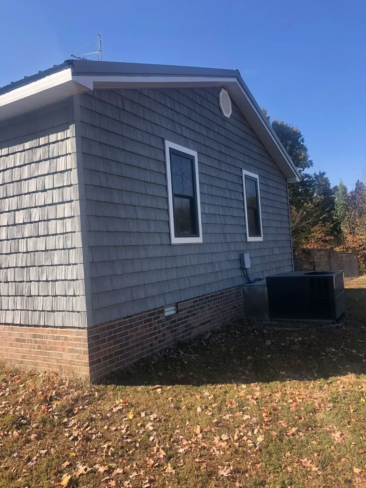 Gray shingle-sided house with white-trimmed windows and brick foundation, set against a blue sky.