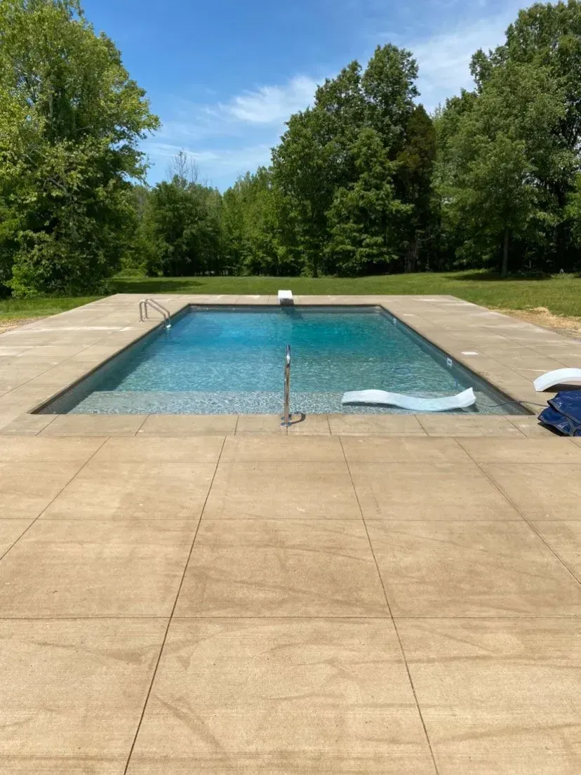 Rectangular pool with turquoise water, surrounded by concrete, in a grassy setting with trees under a blue sky.