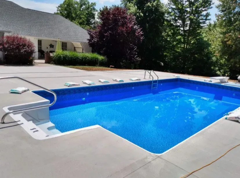 Blue, rectangular swimming pool surrounded by concrete, next to a white house with a dark roof and lush trees.