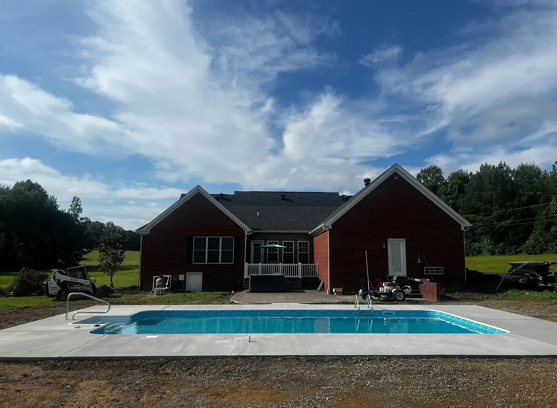 A backyard pool in front of a red house, under a partly cloudy blue sky.