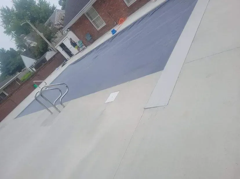 Pool covered with a blue tarp, with a ladder, set in a concrete patio next to a brick house.