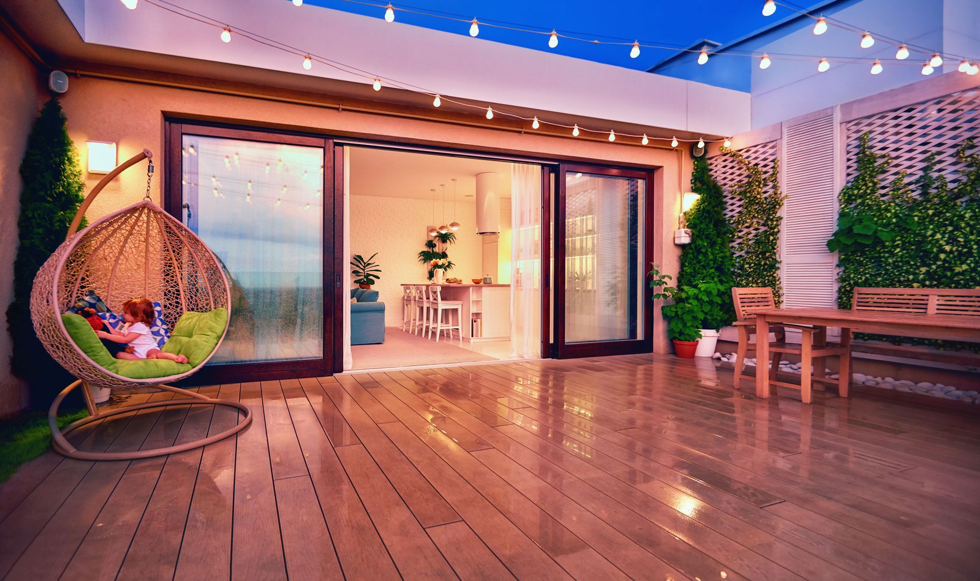 Rooftop patio with hanging chair, open doors to interior, string lights, wooden deck and table, green plants.