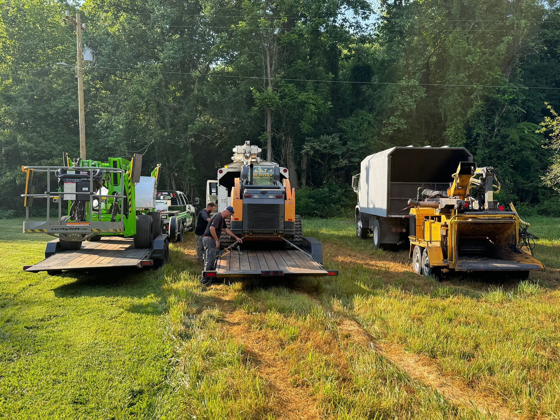 Three pieces of tree removal equipment on trailers parked on grass: a lift, a skid steer, and a wood chipper.