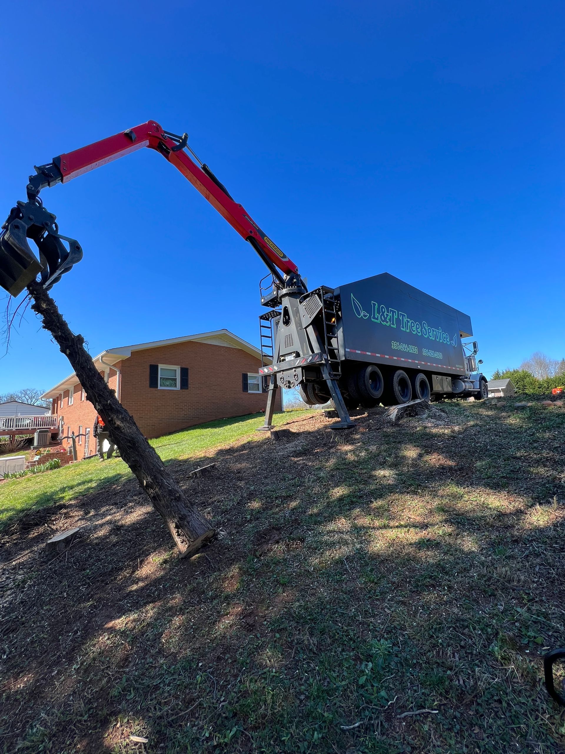 A large truck with a crane grinding a tree stump on a hillside under a blue sky.