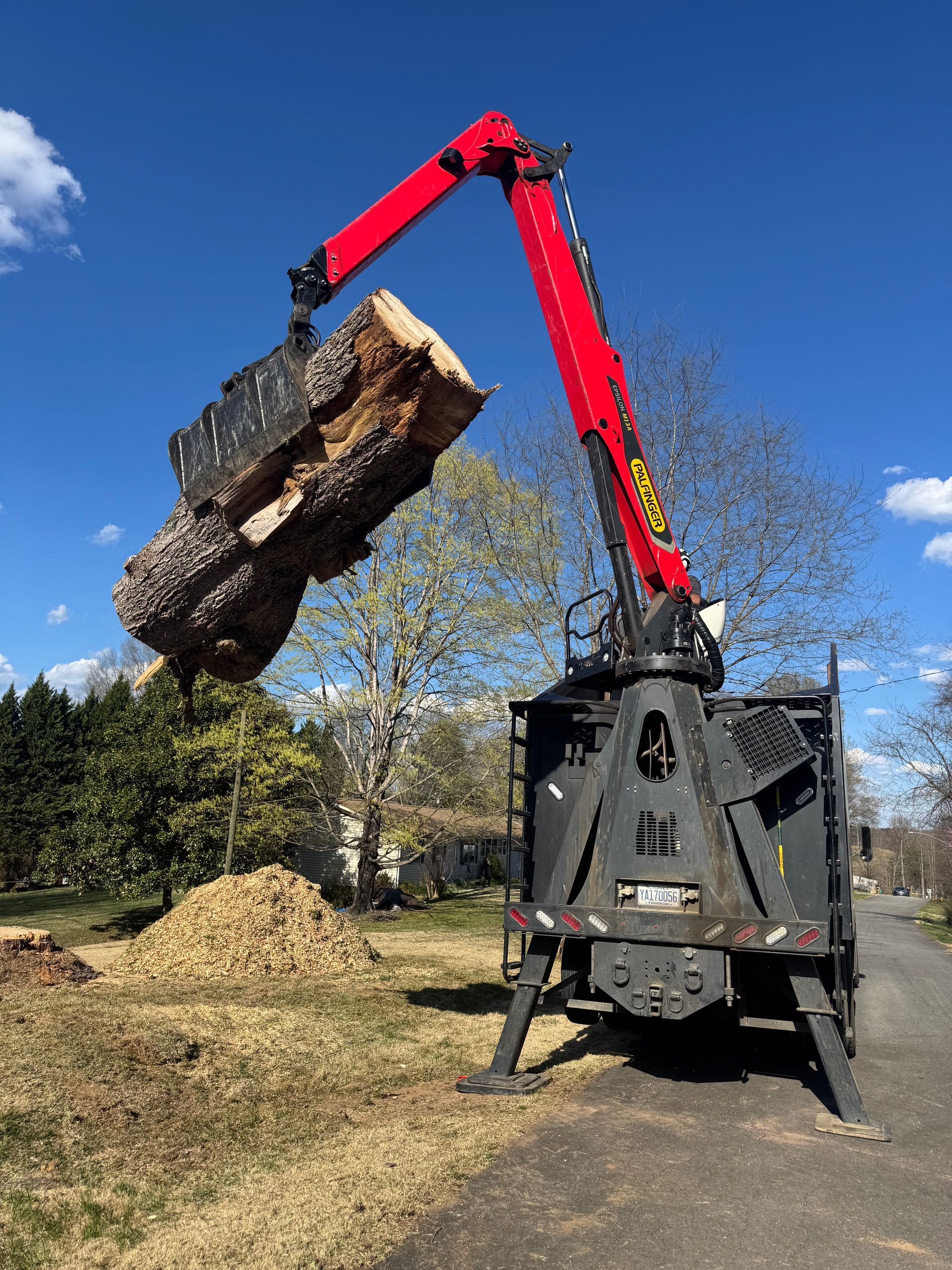 A red and black crane lifting a large log, with wood chips in the foreground, against a blue sky.