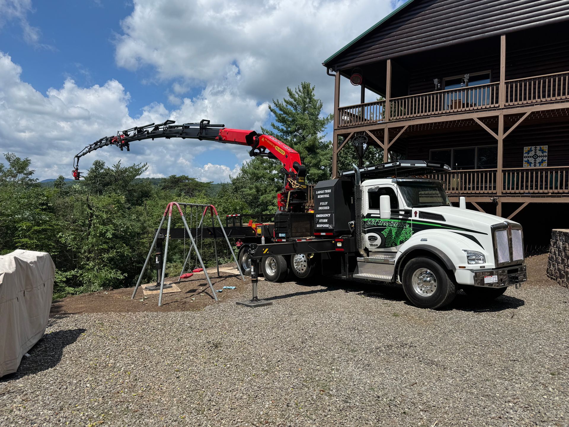 A crane truck parked near a house with its boom extended, working on the surrounding landscape under a partly cloudy sky.