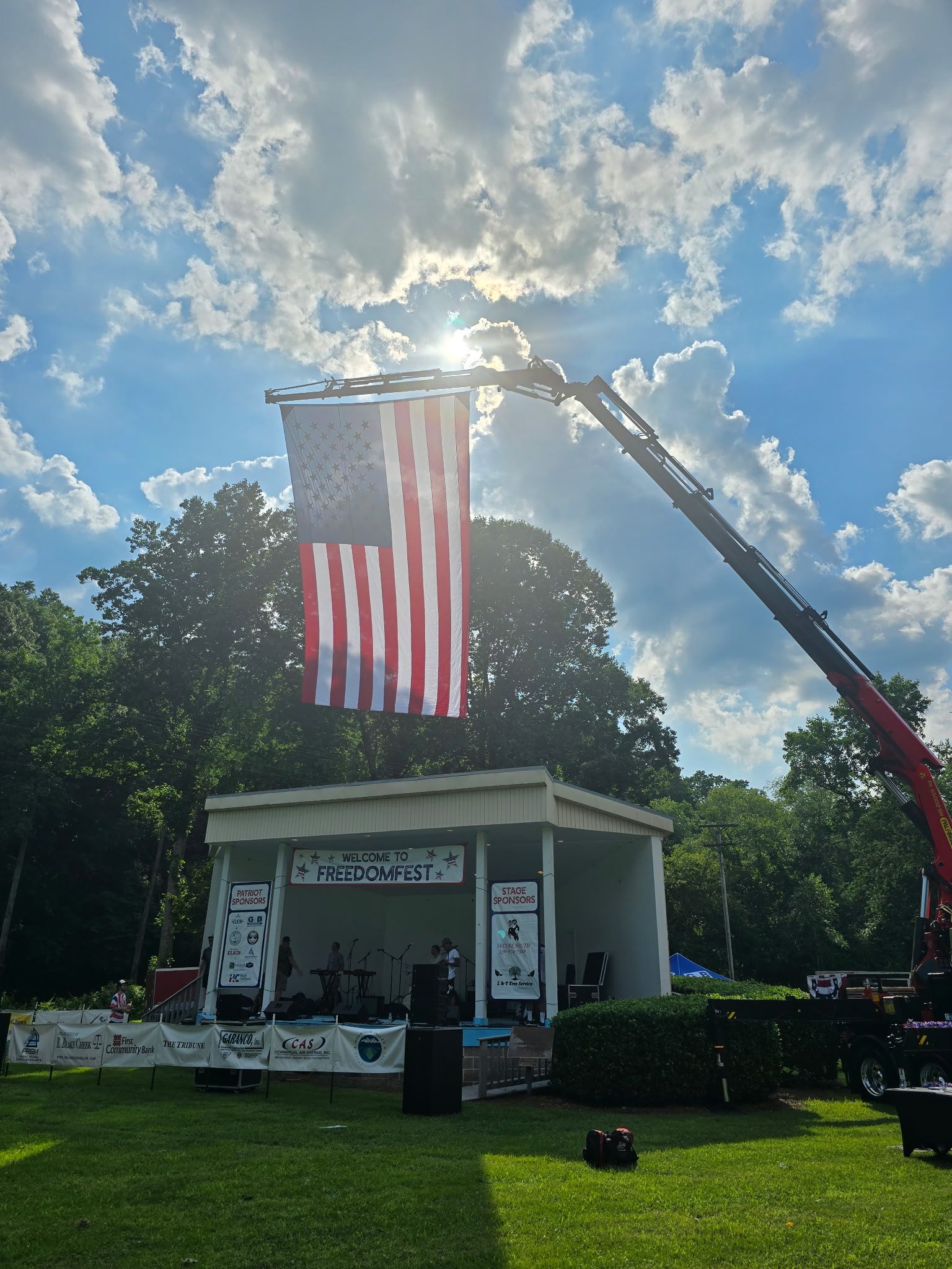 An American flag suspended by a crane over a stage in a park on a sunny day.