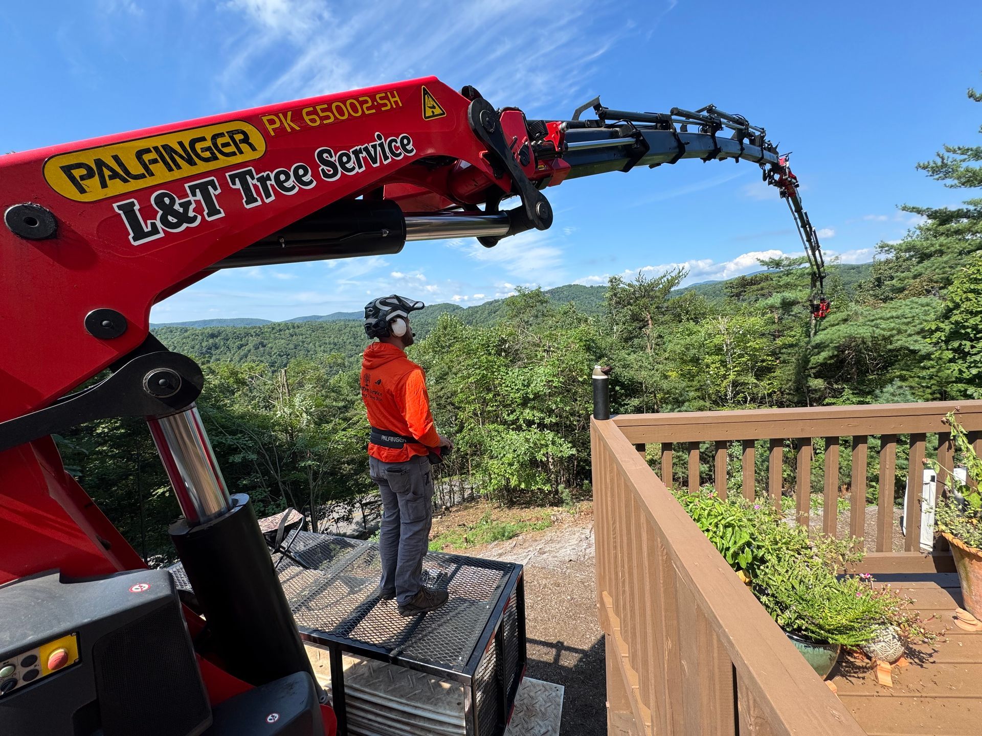 Man in safety gear operates a tree trimming crane on a deck overlooking a forest.