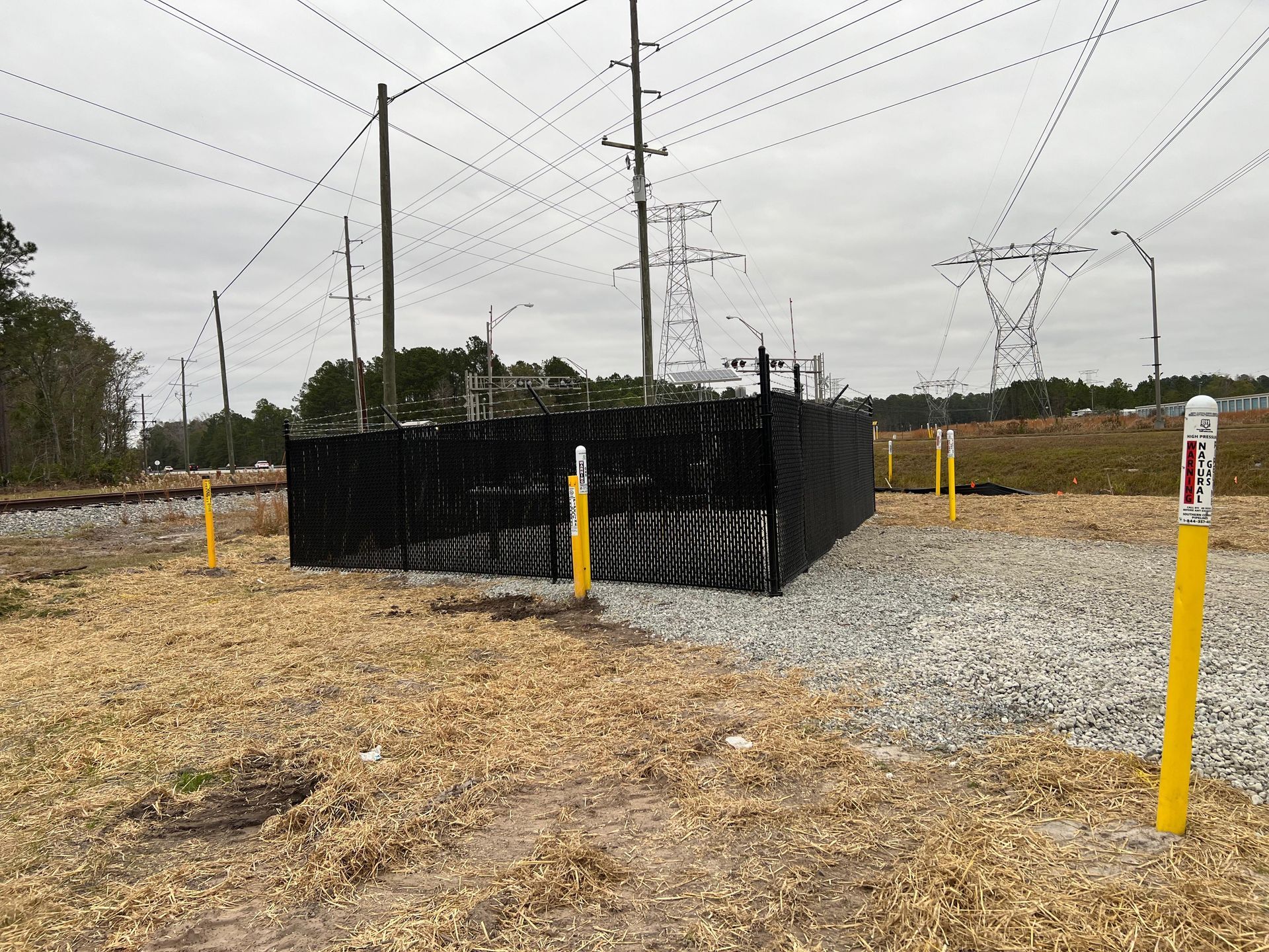 Black fence with yellow posts in a gravel area near power lines and railroad tracks under an overcast sky.
