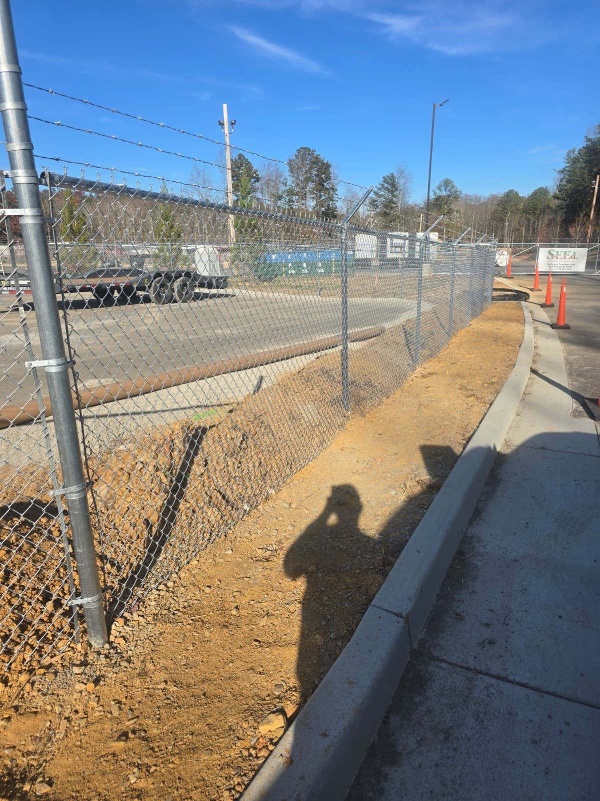 A person's shadow cast on freshly tilled dirt next to a curb and chain-link fence on a sunny day.