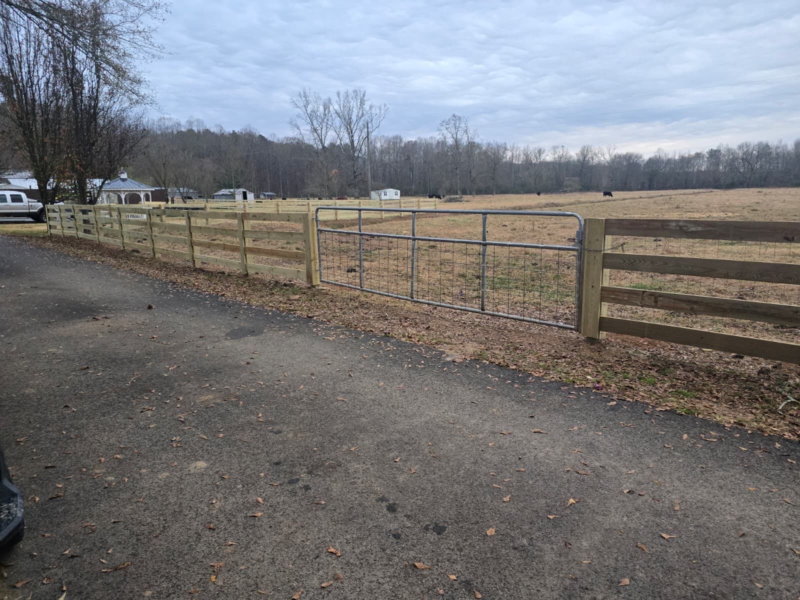 Wooden fence with metal gate in front of a field and trees, under an overcast sky.