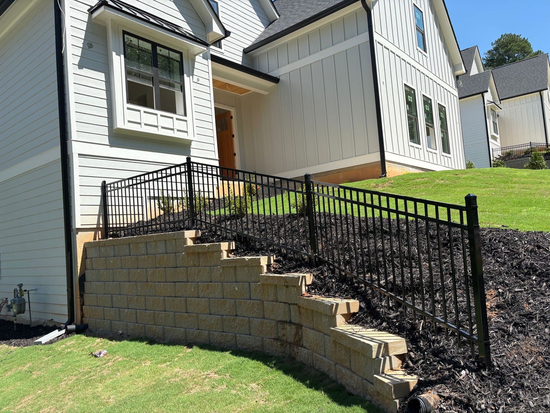 Exterior view of a house with a retaining wall, black fence, and grass.