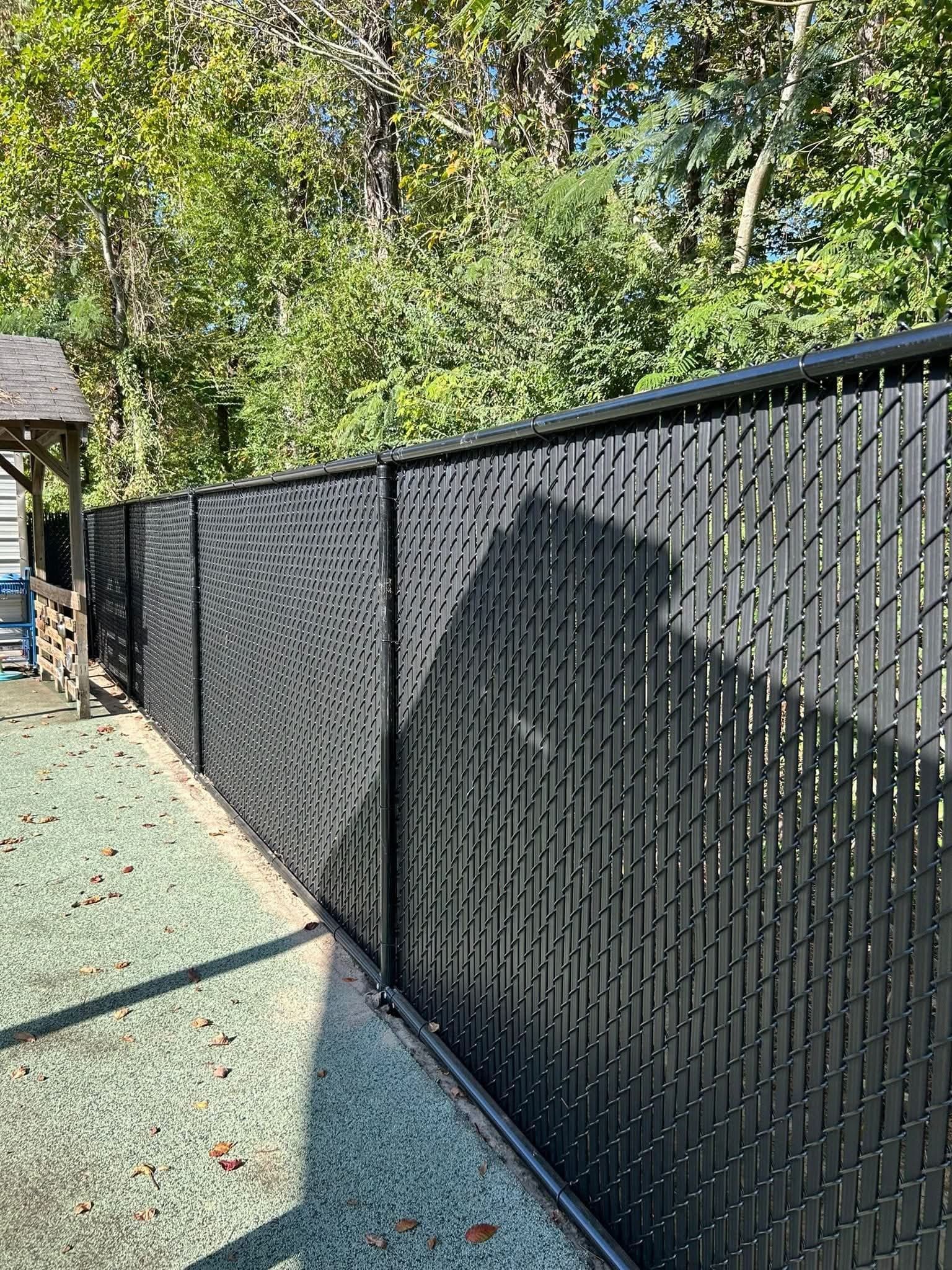 Black chain-link fence with privacy slats, in front of trees. Outdoors, bright sunlight.