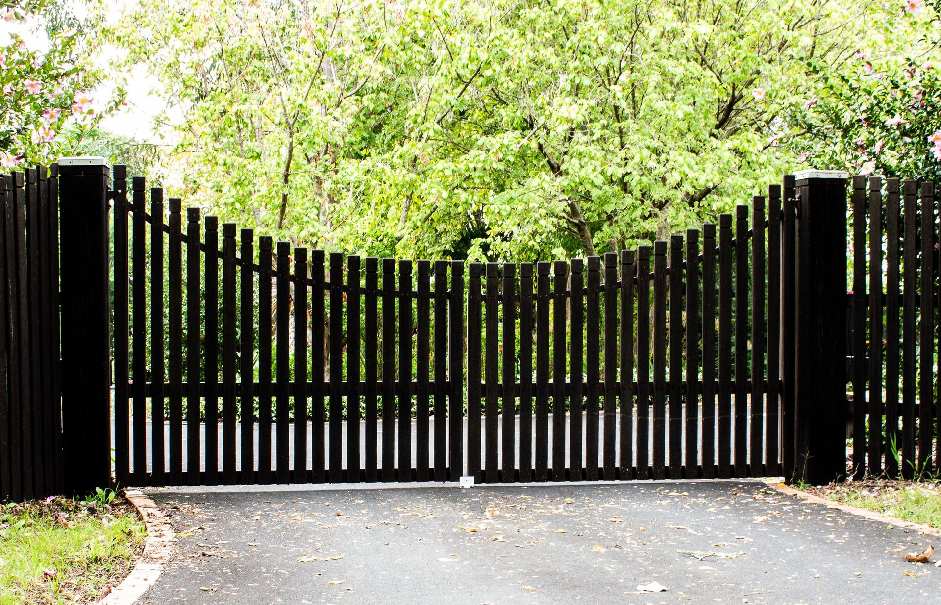Black wooden gate with vertical slats, set in a paved driveway, with trees in the background.
