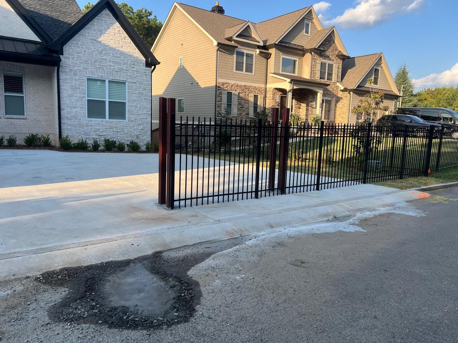 Black metal fence with open gate on a concrete driveway in front of two-story houses.