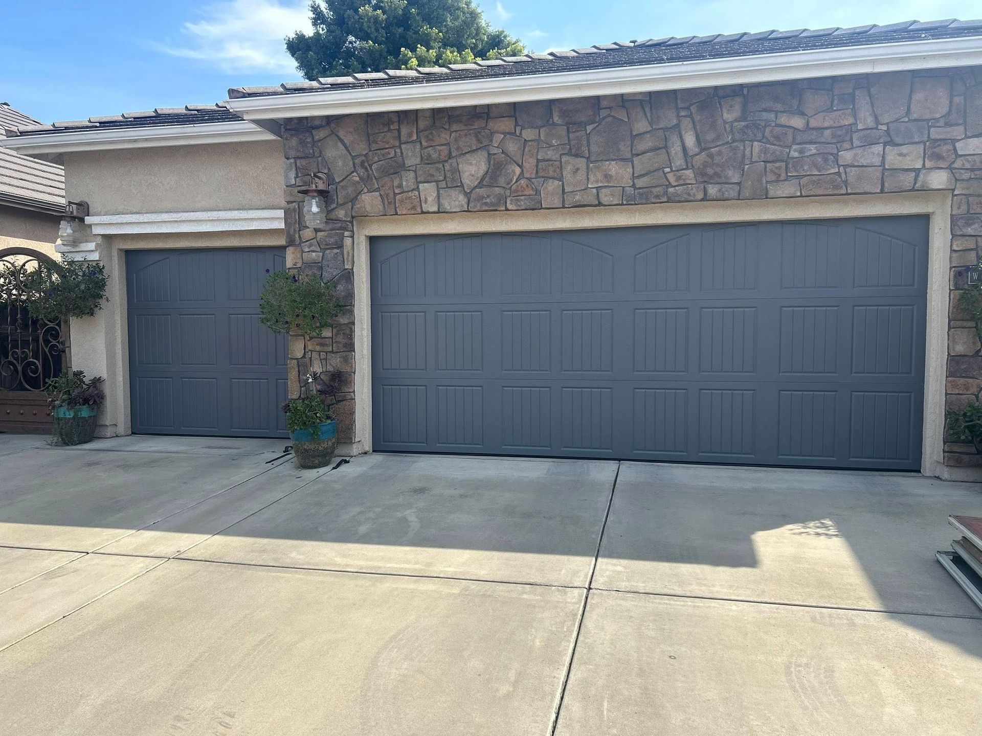 Two gray garage doors on a stone-front house with a concrete driveway and blue sky above