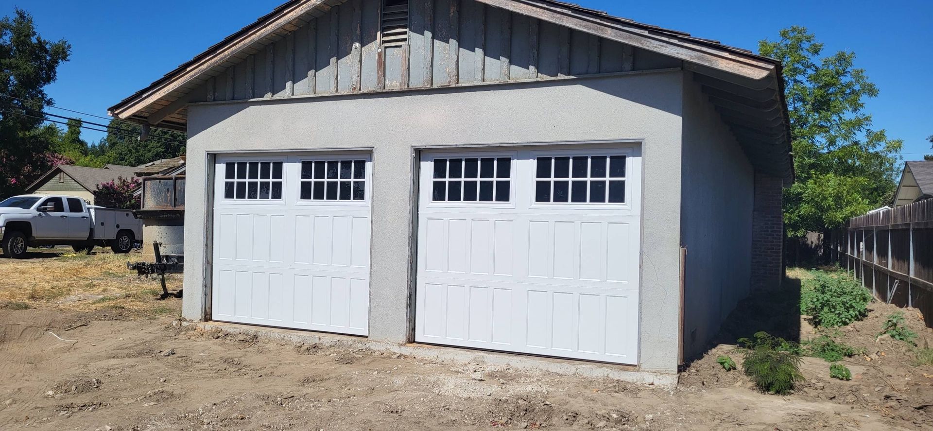 Detached gray two-car garage with white doors on a dirt lot under a clear blue sky