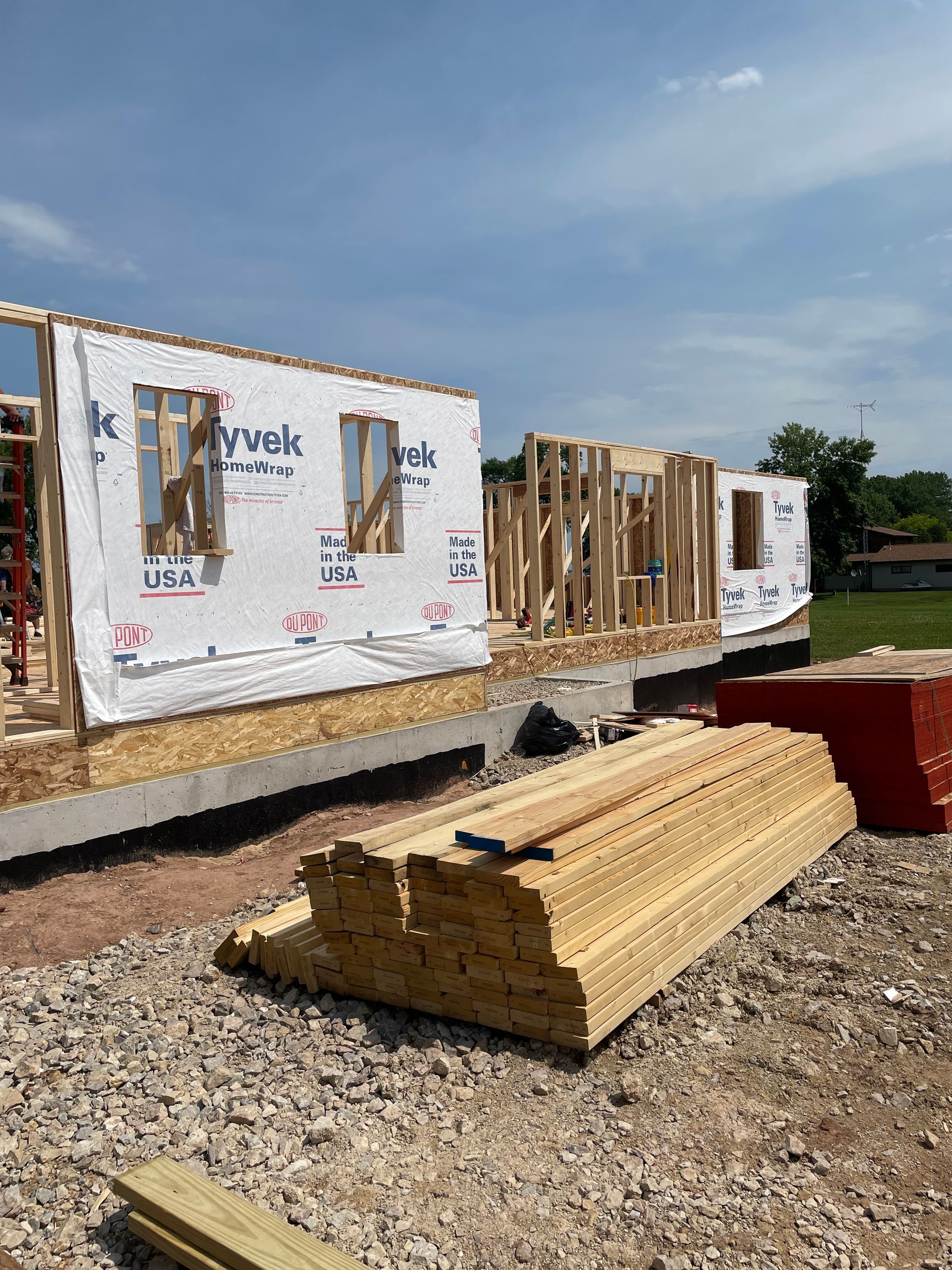 Construction of a house: Wooden framework with Tyvek wrap, unfinished walls, and stacks of lumber on a gravel lot under a blue sky.