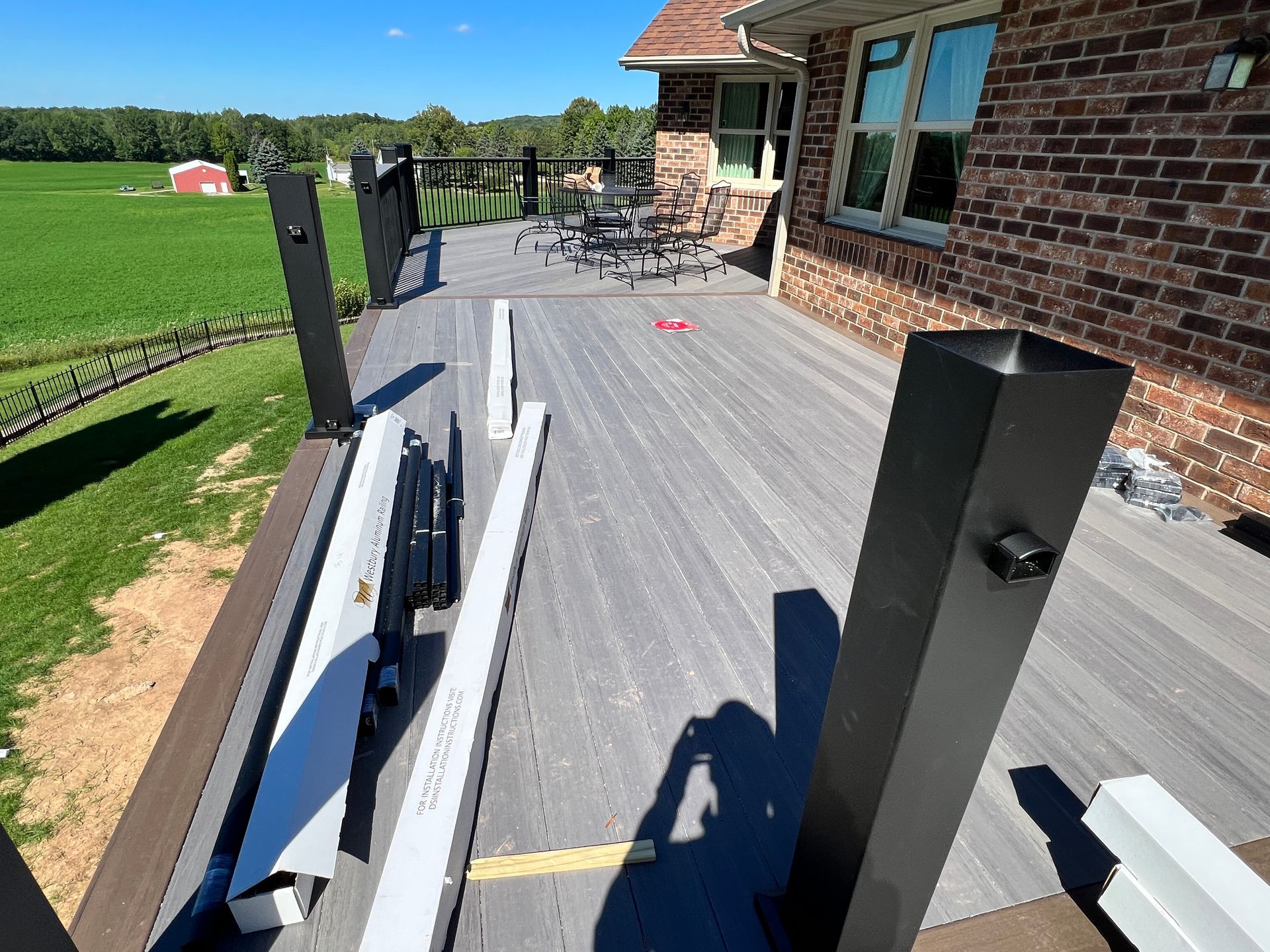 Deck under construction next to a brick house, featuring gray composite decking, black and white railing components, and a field in the background.
