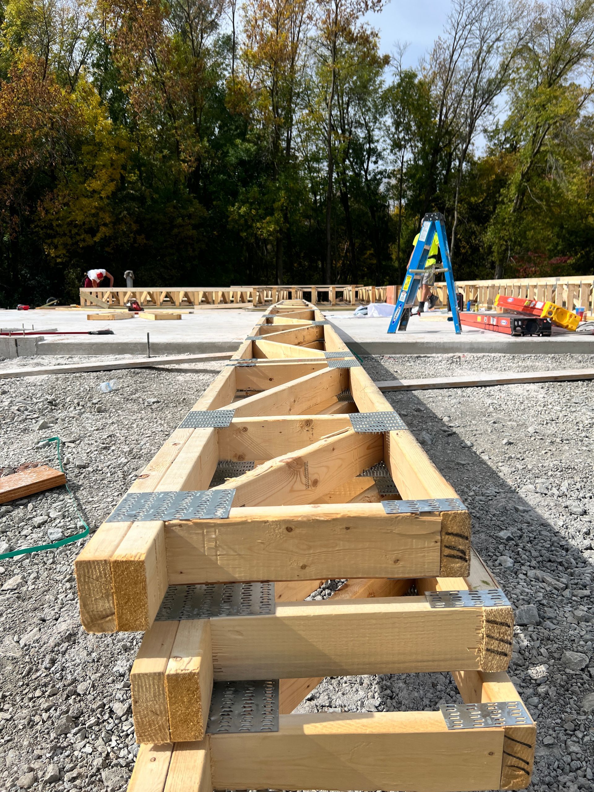 Wooden trusses being assembled on a gravel foundation, likely for a building construction. A blue ladder and other construction materials are in the background.