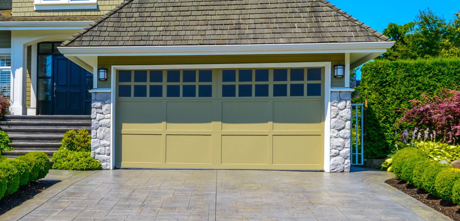 A beige garage door with windows, surrounded by stone pillars and green landscaping, with a concrete driveway.