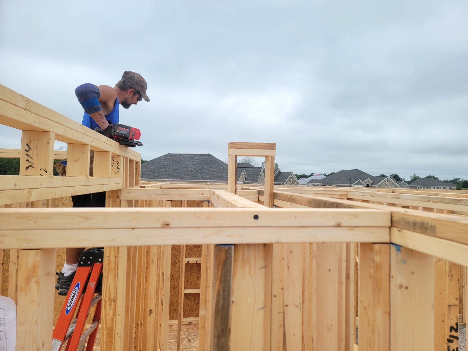 A construction worker uses a power tool on a wooden frame, standing on a ladder at a building site under a cloudy sky.
